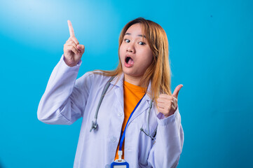 Asian female doctor smiling cheerfully while pointing up with one hand and giving a thumbs up with the other, wearing a white coat and stethoscope on a bright blue background