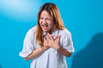Asian woman clutching her chest in discomfort, eyes closed and teeth clenched, expressing intense pain or distress, wearing white shirt with vibrant blue background in studio setting