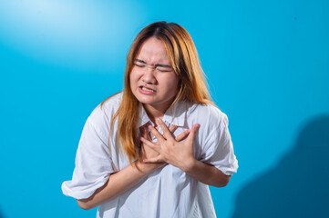 Asian woman clutching her chest in discomfort, eyes closed and teeth clenched, expressing intense pain or distress, wearing white shirt with vibrant blue background in studio setting