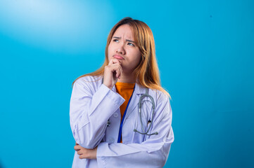 Pensive Asian female doctor in white coat and orange shirt resting chin on hand with thoughtful expression, looking up while standing against bright blue background in studio setting