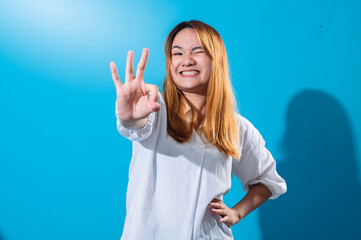 Asian woman with long straight hair in a white shirt smiles brightly while making an OK hand gesture with one hand and winking playfully against a plain blue background in studio lighting
