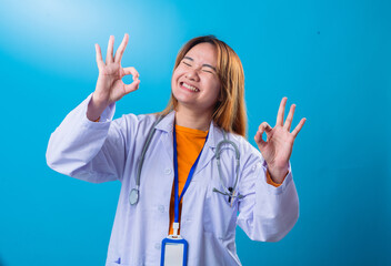 Energetic Asian female doctor in white coat smiling with mouth open, raising both hands in OK gesture, wearing stethoscope, orange shirt, and blue lanyard against blue studio background