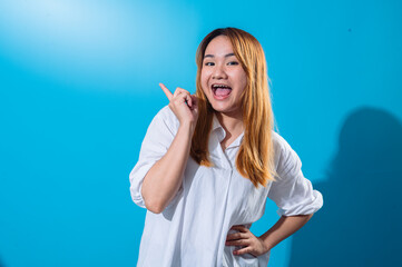 Asian woman in white shirt smiling brightly while pointing upward with index finger on one hand and other hand on her waist, suggesting an idea or highlight, against a solid blue background