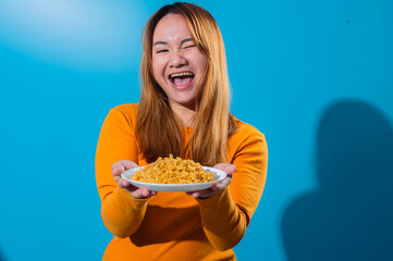 Happy Asian woman in orange shirt smiling and winking while holding a plate of instant noodles with...