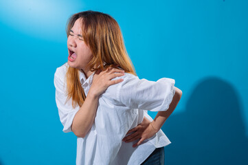 Asian woman wincing in pain while holding her shoulder and back, wearing a white shirt with eyes closed, conveying discomfort or injury, set against a bright blue studio background