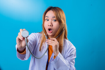 Smiling Asian female doctor wearing white coat and orange shirt, holding stethoscope chestpiece forward with eyes closed in joyful expression against bright blue background
