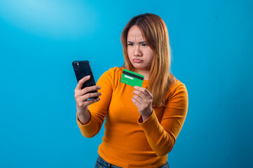 Asian woman in orange shirt looks confused and frustrated while checking her phone and holding a green credit card, against a solid blue background in a studio shot