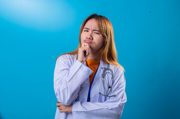 Pensive Asian female doctor in white coat and orange shirt resting chin on hand with thoughtful expression, looking up while standing against bright blue background in studio setting