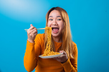 Asian woman in an orange sweater looks surprised while lifting a bunch of instant noodles with chopsticks, holding a white plate, set against a bright blue background