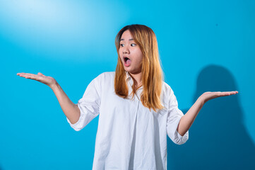 Asian woman with long straight hair in white shirt shows shocked expression with open mouth while raising both hands sideways, standing in front of solid blue background in studio lighting