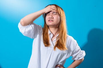Asian woman in white shirt expressing discomfort or frustration, pressing her palm against her forehead with eyes closed, standing against a vibrant blue background in a dramatic studio pose