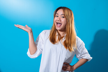 Asian woman with long straight hair in white shirt smiling while holding one hand open to the side as if presenting or showing something, standing confidently against a solid blue studio background