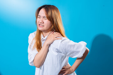 Asian woman wincing in pain while holding her shoulder and back, wearing a white shirt with eyes closed, conveying discomfort or injury, set against a bright blue studio background