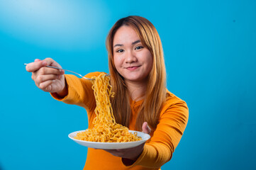 Asian woman in orange sweater smiling and offering a forkful of instant noodles towards the camera...