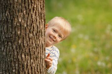 A cheerful blonde-haired boy with bright blue eyes playfully peeks out from behind a textured tree trunk, offering a sweet, engaging smile directly at the viewer.