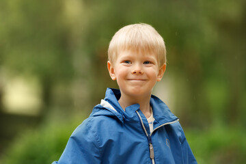 A portrait captures a charming young boy with bright blonde hair and a sweet, slightly mischievous smile. He is wearing a blue hooded jacket, suggesting he's enjoying time outdoors