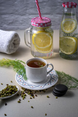 Cup of fennel infusion with fresh herbs, lemon water in glass jars, and a towel on a spa inspired surface. Natural wellness and healthy lifestyle concept
