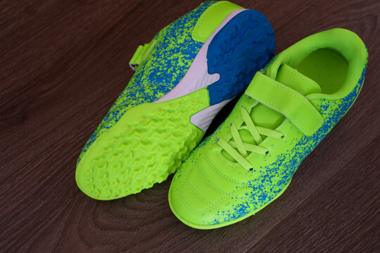 Bright lime green football boots with blue speckles lie on a wooden surface, showing their readiness for a game. Their modern design and sporty look are ideal for themes of sports, childhood, and acti