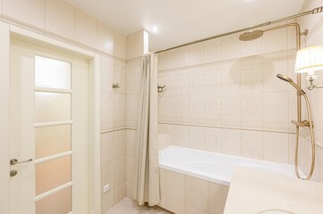 Bright bathroom with beige tiles, a white tub behind a beige curtain, and a shower with golden fixtures. A partially visible door on the left
