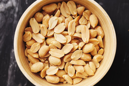 Salted peanuts in wooden bowl. Roasted snack on black marble table. Crunchy golden nuts close-up. Delicious source of protein and energy. Perfect for party, movie night or healthy lunch break.