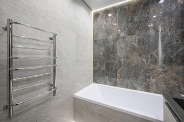 modern bathroom featuring light gray stone tiles, a heated towel rack, and a white bathtub accented...