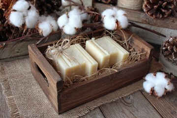 Wooden tray holding two bars of creamy-colored soap, nestled in straw, surrounded by cotton bolls and pine cones