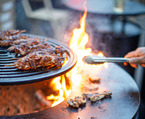 Grilling skewered meat over open fire on a round outdoor barbecue. Juicy meat sizzles on flames as someone uses tongs to turn pieces. Outdoor cooking at its finest.