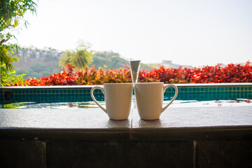 Two coffee mugs placed together with a blurred background of a serene swimming pool, symbolizing a romantic getaway or honeymoon vacation in India. couple holidays, tropical retreats, or leisure