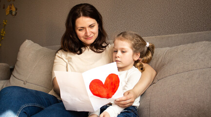 Young girl with light hair hugs mom tightly while holding up her childish drawing. Pure mother-daughter love.