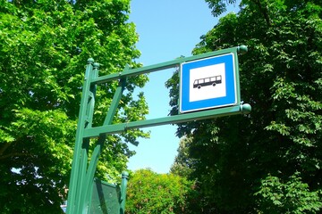 A bus stop sign with a blue frame on a green-painted metal structure, behind it tall, leafy trees...