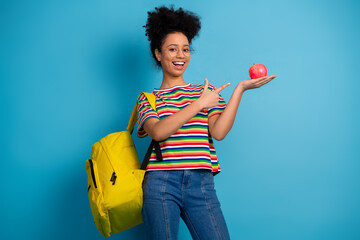 Young woman in colorful striped shirt holding red apple and pointing while smiling against a bright...
