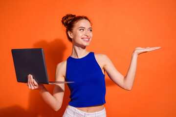 Happy Young Woman Showcasing Laptop with Orange Background in Casual Stylish Outfit Holding Hand...
