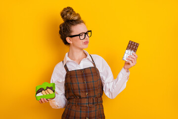 Cute student deciding between chocolate and cookies on yellow background in academic outfit