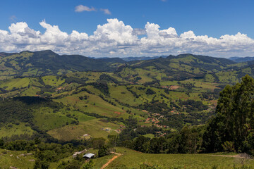 A scenic landscape of rolling green hills and mountains captured in Extrema, Minas Gerais, Brazil. The image shows lush pastureland, forest patches, and dramatic cloud formations against a deep blue