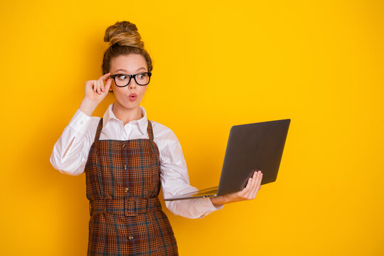 Young woman in glasses holding a laptop on yellow background, expressing curiosity, representing technology and learning