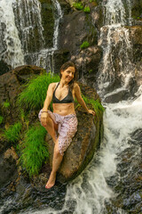 A woman sitting on a rock on the bank of a river or stream, in the middle of a dense forest. Its relaxed pose and natural light filtering through the trees convey a sense of peace, freshness