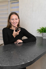 Smiling young woman recording a voice message on her smartphone while comfortably leaning against the kitchen counter in her stylish modern apartment, enjoying a casual moment of connection.