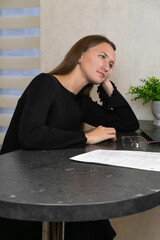 Young businesswoman in black clothes sitting at table with documents, glasses and laptop, leaning on her hand and looking away, feeling tired and bored. Vertical photo