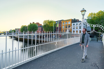 A man walks across Dublin’s iconic Ha’penny Bridge at sunset, with colorful riverside buildings and calm waters in the background.
