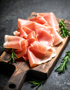Thin slices of prosciutto on wooden board with rosemary. Tasty food.