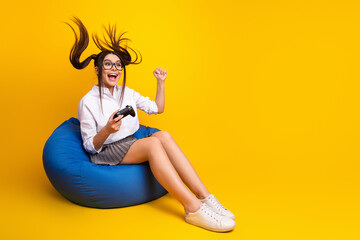 Excited young woman playing video games on bean bag with dynamic action against a vibrant yellow...