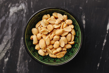 Salted peanuts in ceramic bowl. Healthy protein snack. Crunchy appetizer on black marble background. Natural texture and detail. Tasty vegetarian food. Simple rustic kitchen scene top view.