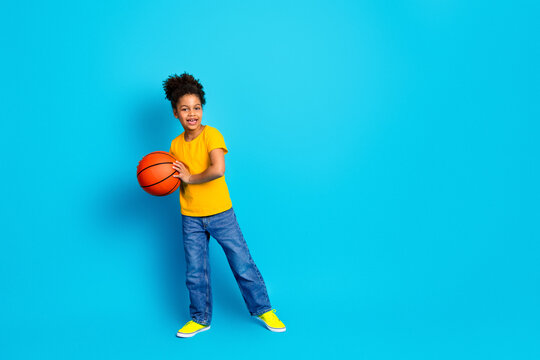 Young girl holding basketball playfully standing against bright blue background, wearing yellow t-shirt and jeans, enjoying leisure time.