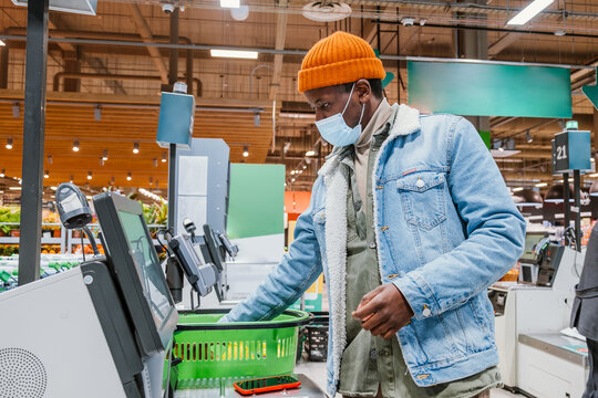 Young black man wearing a protective face mask is using a self-checkout counter in a supermarket, placing his shopping basket on the counter and holding his credit card