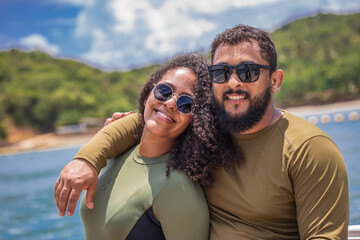 A cheerful couple poses together outdoors near a body of water, both wearing sunglasses and sun-protective clothing. Their relaxed expressions and close posture convey a sense of joy and connection. 