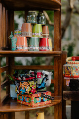 Artfully stacked and painted cups and kettles sit on wooden shelves, showcasing vibrant colors and unique designs in an outdoor setting.