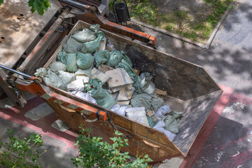 Top view of a construction dumpster filled with green bags containing building debris and rubble, surrounded by trees casting shadows on a sunny day