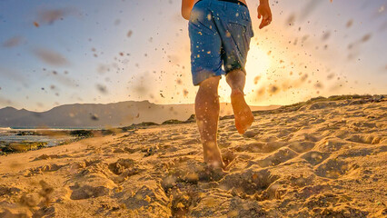 LOW ANGLE VIEW, LENS FLARE: Fit young man in blue shorts runs barefoot across sandy beach at sunrise, kicking up sand in his path. Low sun casts a golden glow while he is already on his morning run.