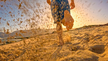 LOW ANGLE VIEW, LENS FLARE: Athletic man in blue shorts runs barefoot along sandy ocean beach in golden light. He is on an early morning run and enjoys relaxing and active holidays at Lanzarote island