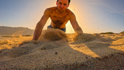 LOW ANGLE VIEW: Clumsy man stumbles and falls into sand while running beach in early morning. Sand flies in all directions in golden light while young man hits the sandy ground after spectacular fall.